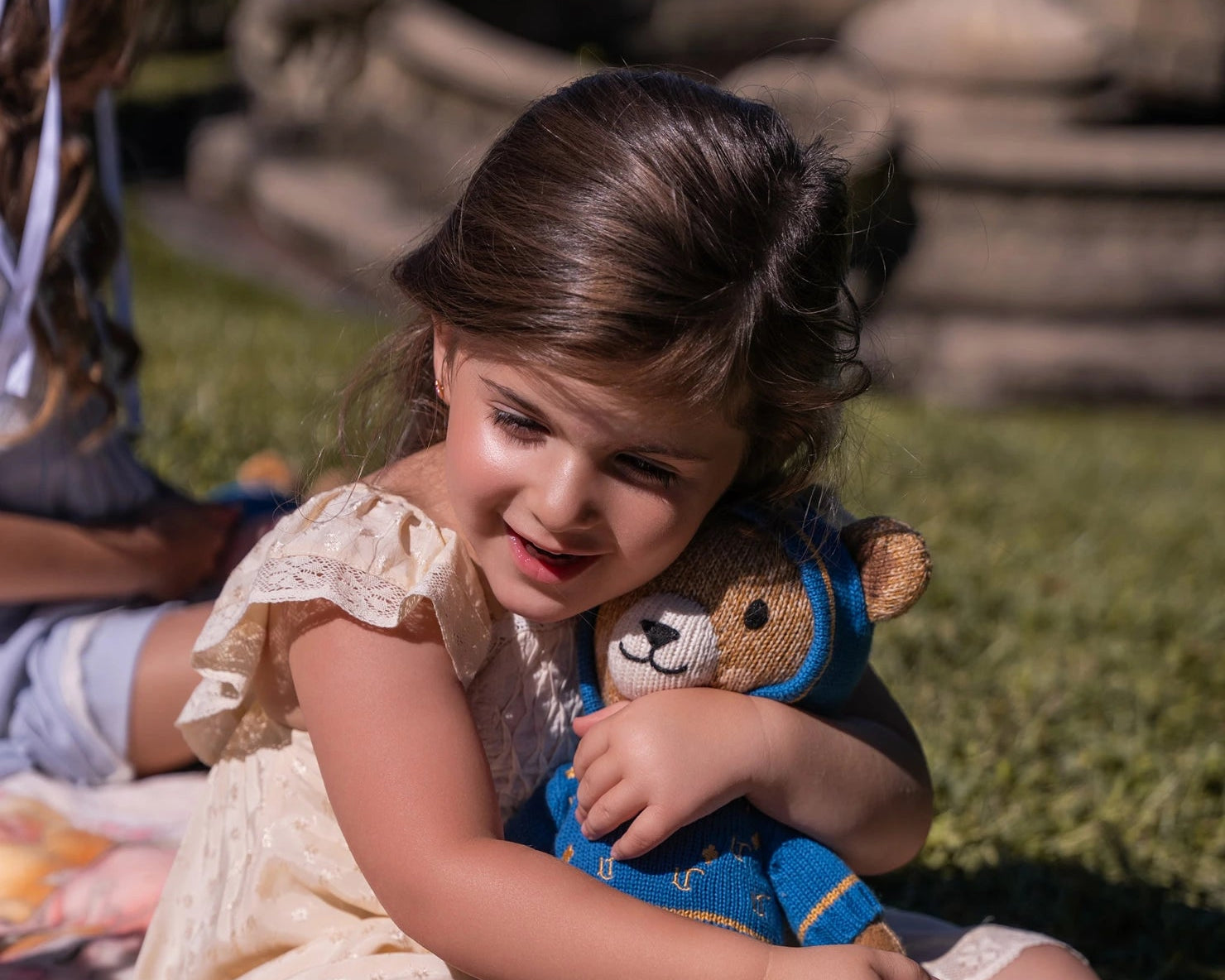 Young girl sitting on grass holding a teddy bear, wearing a yellow dress with white lace trim.